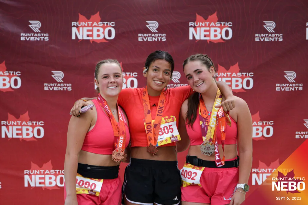 Three smiling female finishers at Runtastic NEBO with award medals, in front of a red photo backdrop